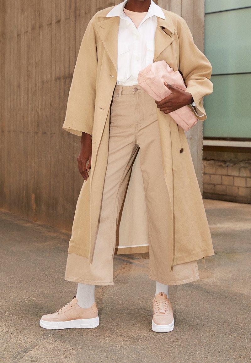 Beige trench coat, valkoinen paita, vaaleanbeige leveälahkeiset housut, vaaleanpunaiset tossut valkoisilla pohjilla, pitäen vaaleanpunaista clutch-laukkua. Sileä kangas, hyvin muotoiltu design.