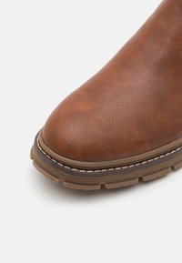 Botte en cuir marron avec un bout arrondi, une surface texturée et une semelle en caoutchouc épaisse. Présente des coutures contrastées autour de la base.