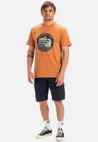 Orange cotton t-shirt featuring a circular graphic print of a wave and sun, paired with navy cargo shorts and black sneakers.