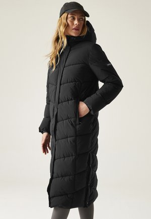 Kobieta ubrana w długi czarny pikowany płaszcz puchowy z kapturem i zatrzaskami po bokach, w czarnej czapce bejsbolowej, stojąca na jednolitym tle.