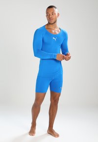Blå atletisk bodystocking med lange ærmer og shorts. Fremstillet af glat, strækbart materiale. Har et lille hvidt logo på brystet.