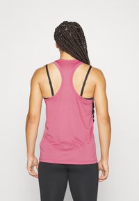 Rosa atletisk linne med racerback-design, slät textur och svarta justerbara remmar. Den nedre fållen är något böjd. Svarta leggings är på.