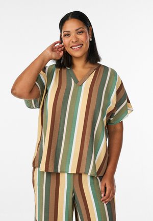 Nasmejana ženska, oblečena v ohlapno kratko majico in hlače z navpičnimi črtami v zelenih, rjavih, rumenih in bež odtenkih.