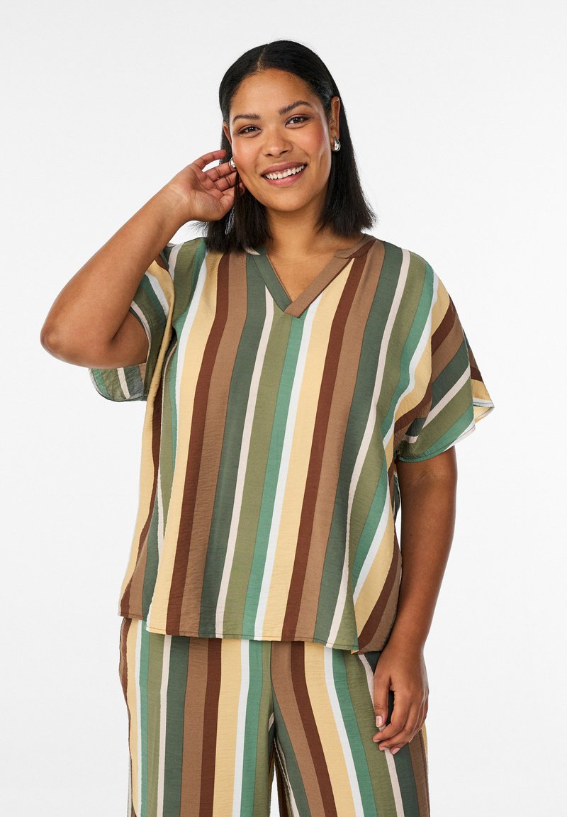 Lächelnde Frau trägt ein lockeres, kurzärmeliges Oberteil und eine Hose mit vertikalen Streifen in Grün-, Braun-, Gelb- und Beigetönen.