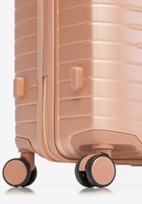 Primer plano de una esquina de una maleta rígida color oro rosa que muestra ruedas giratorias dobles y una cremallera parcialmente visible.
