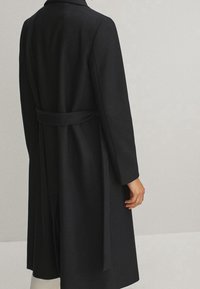 Manteau en laine noire au design ajusté, manches longues et ceinture à la taille. Présente un col cranté et une texture lisse.