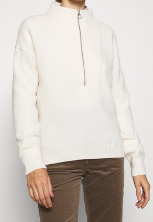 Pull tricoté écru avec col demi-fermeture éclair associé à un pantalon en velours côtelé marron porté par une personne debout devant un fond uni.