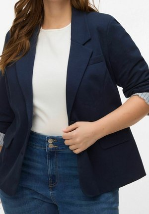 Femme portant un blazer bleu foncé aux manches retroussées, un haut blanc et un jean bleu taille haute, tenant l'avant du blazer d'une main.