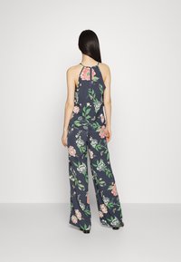 Macacão floral azul-marinho com decote halter, apresentando grandes padrões de rosas cor-de-rosa e verdes, pernas largas e relaxadas, e um detalhe de abertura nas costas.