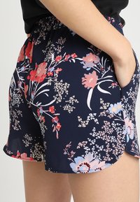 Person klädd i marinblå blommiga shorts med röda, rosa och ljusblå blommor, med en hand avslappnat placerad i fickan mot en enkel bakgrund.