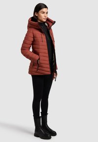 Padded, rust-colored parka with a hood, zip front, and side pockets. Worn over a black top and paired with black leggings and chunky boots.