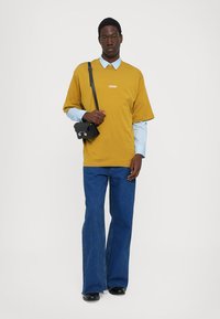 O persoană stă în fața unui fundal alb, purtând un tricou oversized de muștar, o cămașă albastră deschisă dedesubt și pantaloni din denim evazați, având o geantă mică neagră.