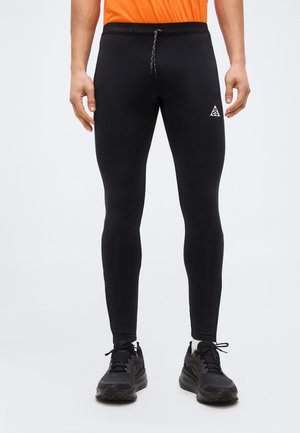 Muž v čiernych športových tesných legínach s logom ACG na stehne, čiernych teniskách a oranžovom tričku, stojaci pred jednoliatym pozadím.