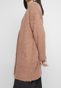 Langer, gestrickter Cardigan in sanftem, staubigem Rosaton. Verfügt über einen lässigen Schnitt, gerippte Textur und abgesenkte Schultern für eine entspannte Silhouette.