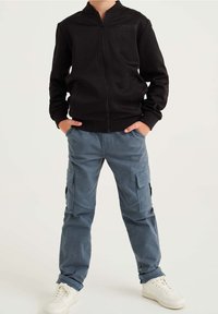 Czarna kurtka zapinana na zamek z prążkowanymi mankietami, w zestawieniu z jasnoniebieskimi spodniami cargo z bocznymi kieszeniami oraz białymi trampkami. Minimalistyczny design.