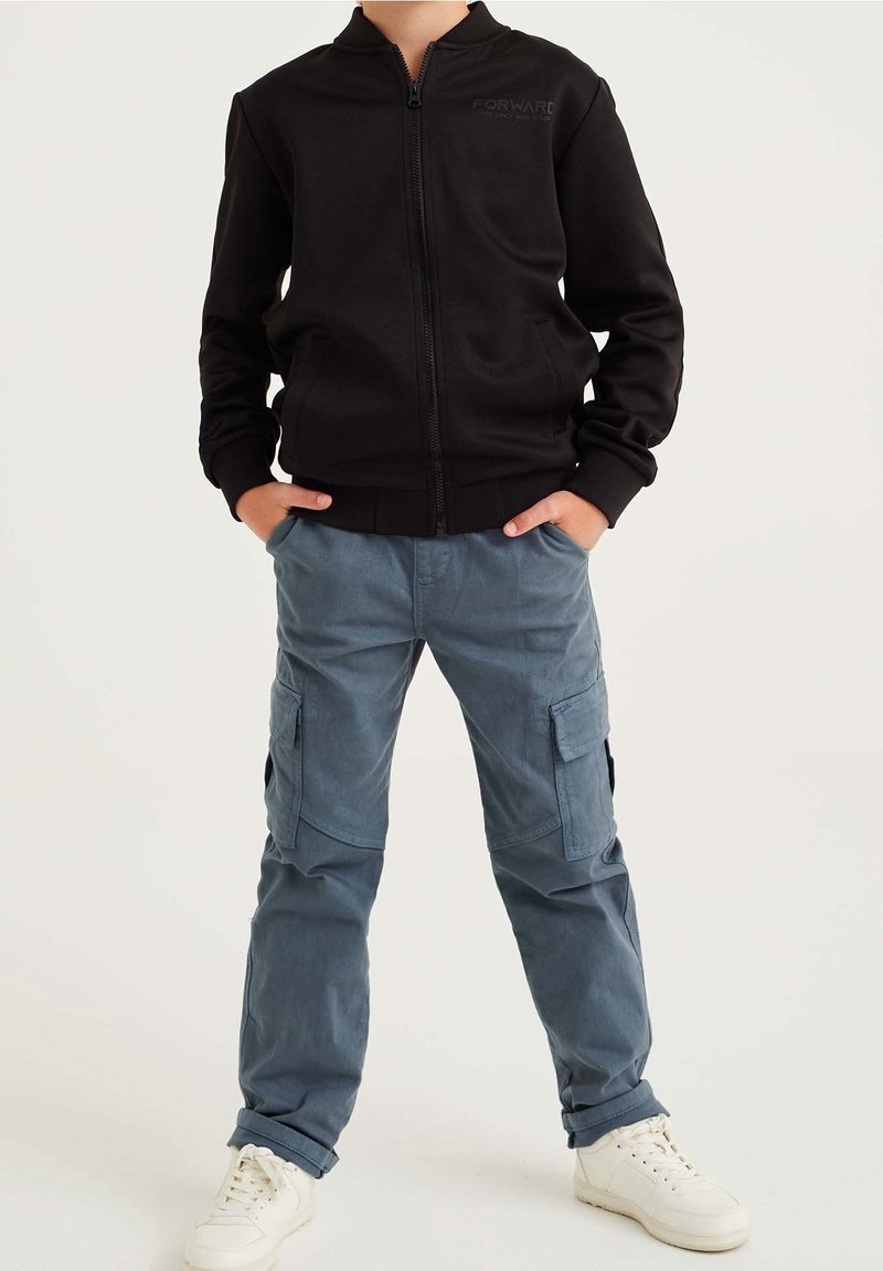 Czarna kurtka zapinana na zamek z prążkowanymi mankietami, w zestawieniu z jasnoniebieskimi spodniami cargo z bocznymi kieszeniami oraz białymi trampkami. Minimalistyczny design.