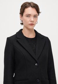 Μαύρο σακάκι από μείγμα μαλλιού, με κανονικό γιακά, κλείσιμο με δύο κουμπιά και πλάγιες τσέπες. Η υφή είναι λεία με διαρθρωμένες ραφές.