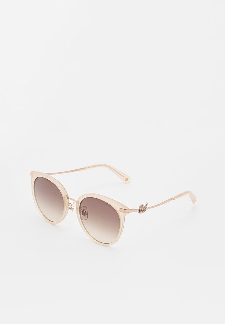 Des lunettes de soleil en forme d'œil de chat avec une teinte brune dégradée. Monture en plastique rose clair avec des branches métalliques dorées fines et un embellissement décoratif sur le côté.