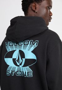 Sort hættetrøje med blåt grafisk design på bagsiden med teksten "VOLCOM" og "STONE", der viser et dristigt geometrisk mønster.