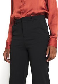 Zwarte op maat gemaakte broek met een gladde textuur en een gestructureerde pasvorm, gecombineerd met een roestkleurige zijden blouse met knoopdetails.