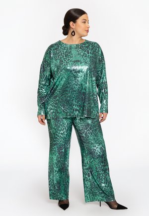 Vrouw in een glanzend groen luipaardprint top en wijde broek, met zwarte puntige hakken en zwarte oorbellen, staand tegen een witte achtergrond.