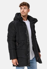 Chaqueta acolchada negra con capucha, cierre de cremallera y botones a presión, bolsillos frontales, tejido texturizado y cordones de contraste.