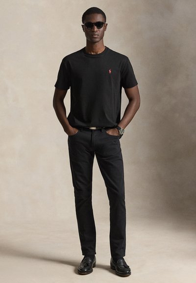 Άνδρας φοράει μαύρα γυαλιά ηλίου, μαύρο μπλουζάκι με κόκκινο λογότυπο, μαύρο παντελόνι, μαύρη ζώνη, μαύρα μοκασίνια με φούντες και ρολόι χειρός, στέκεται με τα χέρια στις τσέπες.