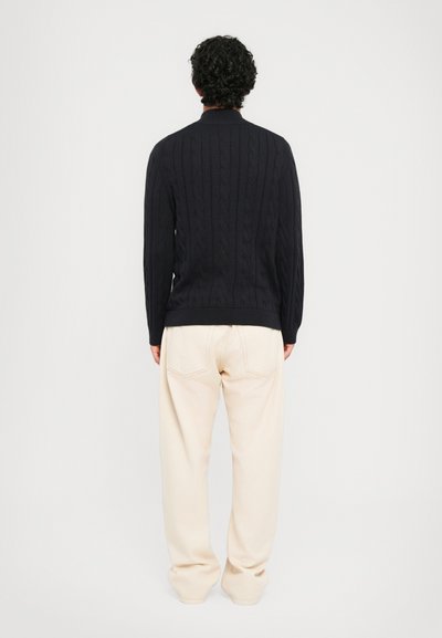 Άνδρας με σγουρά σκούρα μαλλιά, φορώντας ένα μαύρο πλεκτό πουλόβερ με καρώ και φαρδιά μπεζ παντελόνια, που στέκεται στραμμένος μακριά σε έναν απλό λευκό φόντο.