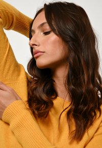 Maglione giallo con polsini a coste, texture morbida. Lunghi capelli scuri e mossi incorniciano il volto, mettendo in risalto i lineamenti delicati e il trucco naturale.