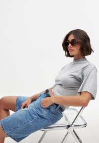 Pelēks īsām piedurknēm tops apvienots ar augsto jostasvietu zilām denima bermudu šortiem. Aksesuāri ietver apaļas bruņurupuča stila saulesbrilles un melnu jostu.