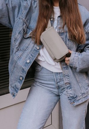Lichtgrijze rechthoekige crossbody tas van glad materiaal, gedragen over een wit shirt, gecombineerd met een lichte spijkerjack en jeans.