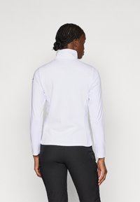 Casaco branco de manga longa com colarinho alto e superfície texturizada, combinado com calças pretas, visto de costas.