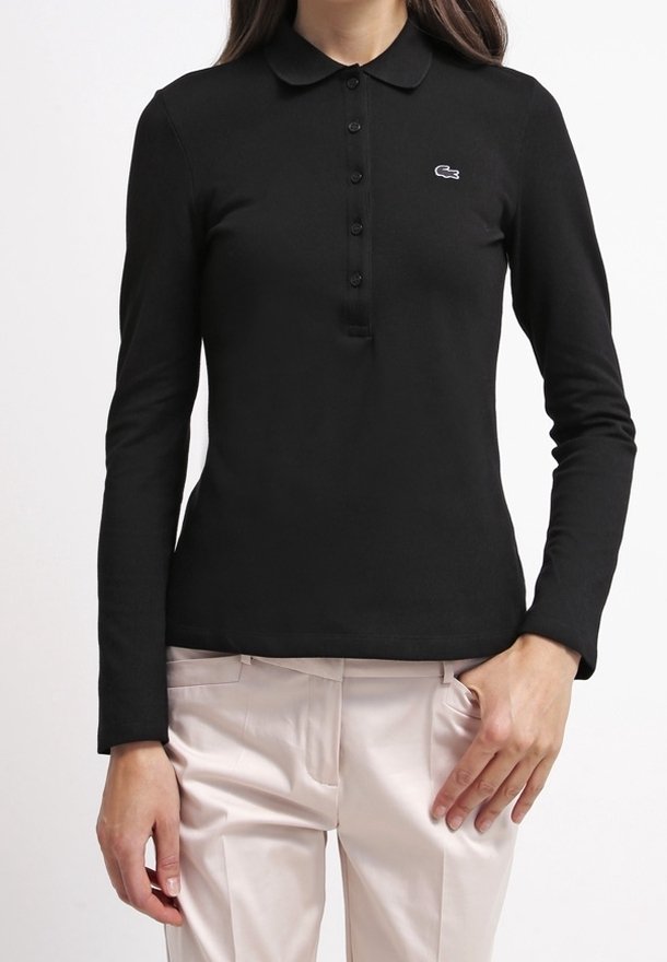 T-shirt polo noir à manches longues avec un col classique, patte de boutonnage et logo Lacoste en blanc. Tissu lisse et ajusté, associé à un pantalon léger.