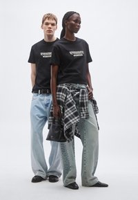 Fekete "Tommy Jeans" logós pólók, világoskék bő fazonú farmernadrágok, és egy fekete-fehér kockás ing, amelyet a derék köré kötve viselnek. Fekete kézitáska és cipők.