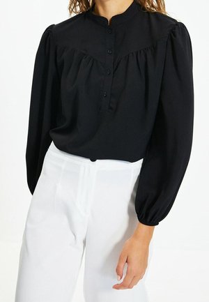 Femme portant un chemisier noir à manches longues avec boutons et col montant associé à un pantalon blanc taille haute sur fond blanc.