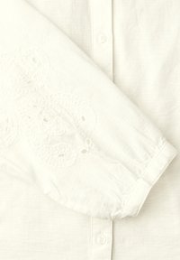 Blouse blanche à manches longues avec fermeture boutonnée sur le devant et broderie florale discrète sur le poignet et le haut du bras.