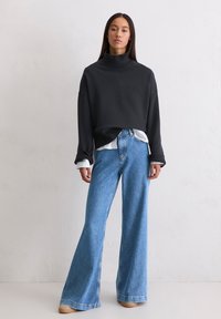 Przesadnie duża czarna bluza z stójką noszona na białej i niebieskiej w paski koszuli, w połączeniu z szerokimi jasnoniebieskimi dżinsami i beżowymi trampkami.