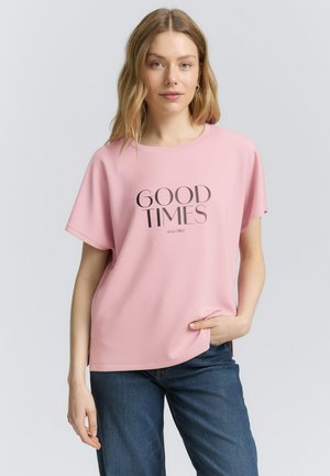 T-shirt rose clair à manches courtes et encolure ronde, avec le texte "GOOD TIMES" en gris foncé et des fentes sur les côtés. Associé à un jean bleu.