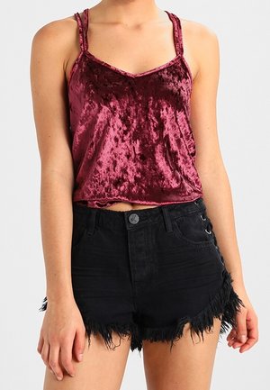 Kobieta ubrana w bezrękawnik z marszczonego aksamitu w kolorze bordowym i wysokiej talii czarne szorty z dżinsu z postrzępionymi brzegami oraz sznurowanymi bokami.