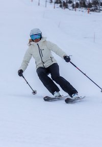 Ski-uitrusting omvat een lichtbeige jas, zwarte broek en blauwe skibril. De skiër draait op een besneeuwde helling met skistokken.