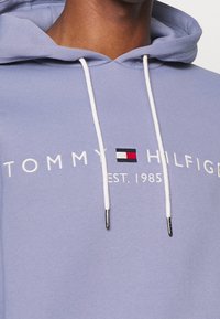 Felpa leggera viola realizzata in tessuto morbido, con cordini bianchi, testo ricamato "TOMMY HILFIGER" e un piccolo logo rosso, bianco e blu.