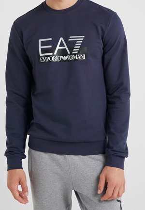 Uomo che indossa una felpa a maniche lunghe blu navy con logo bianco "EA7 Emporio Armani" e pantaloni della tuta grigio chiaro.