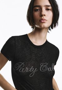 Crna majica kratkih rukava s blago prozirnom teksturom i natpisom "Party Call" u srebrnim zakovicama preko prsa.