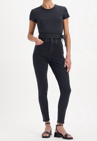 Jeans de mezclilla negros de tiro alto con un ajuste ceñido, que cuentan con cinco bolsillos y cierre de botón. Combinados con una camiseta negra de manga corta ajustada y sandalias.