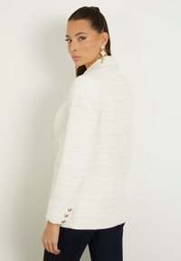 Blazer bianco con texture, dotato di colletto a punta, due tasche frontali e dettagli in bottone dorato sulle maniche. Modello mostrato di spalle.