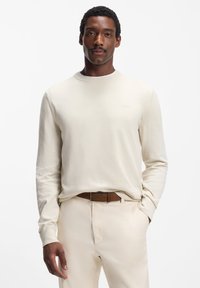 Strickpullover in Cremefarbe mit rundem Ausschnitt, langen Ärmeln und einem dezenten Logo. Getragen über hellbeigen Hosen mit braunem Gürtel.