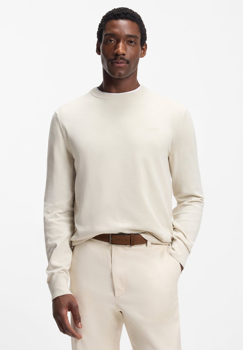 Strickpullover in Cremefarbe mit rundem Ausschnitt, langen Ärmeln und einem dezenten Logo. Getragen über hellbeigen Hosen mit braunem Gürtel.