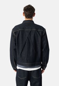 Moški v temno modrem džinsu jopiču in hlačah, prikazan zadaj, s precejšnjo trokutasto pregibno detajl izvajanje na zadnjem delu jopiča.