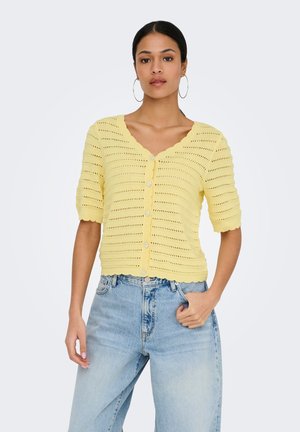 Mujer con cárdigan de punto amarillo de manga corta abotonado y vaqueros de cintura alta azul claro, de pie con una mano en el bolsillo.