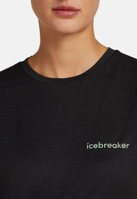 Primo piano della parte inferiore del viso e del collo di una persona che indossa una maglietta nera a girocollo con un piccolo logo verde pallido "icebreaker" sul petto.